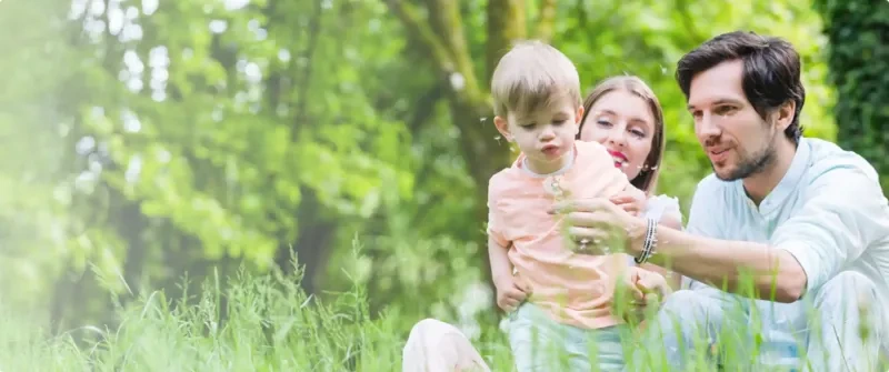 A toddler blows out a dandelion while parents look on.